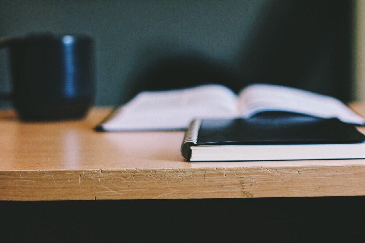 A closed black spiral notebook on a scratched wooden table, with an open book and a dark mug blurred behind it.